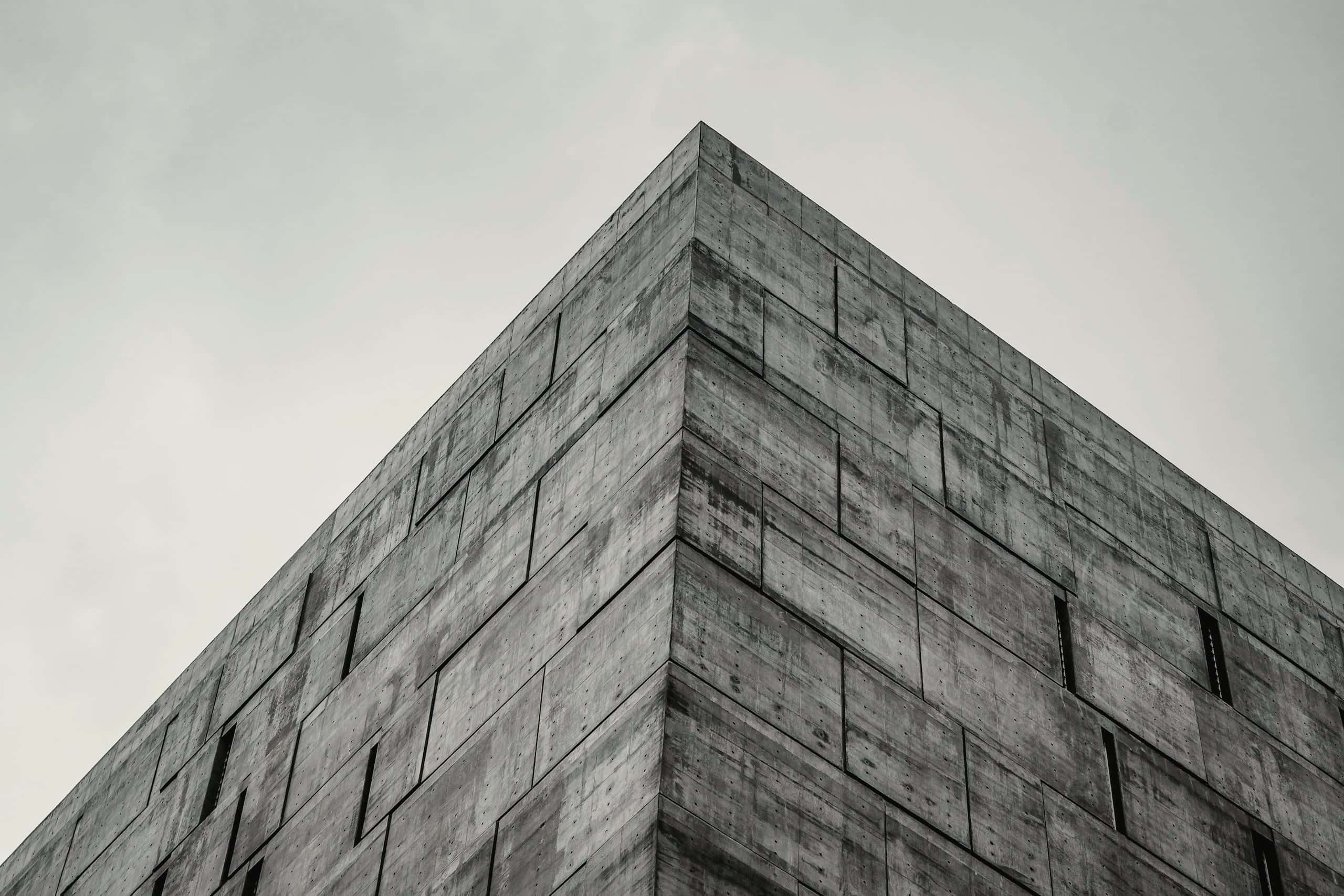 Corner of a board-marked concrete volume rising against an overcast sky, narrow window slots set flush in the wall.