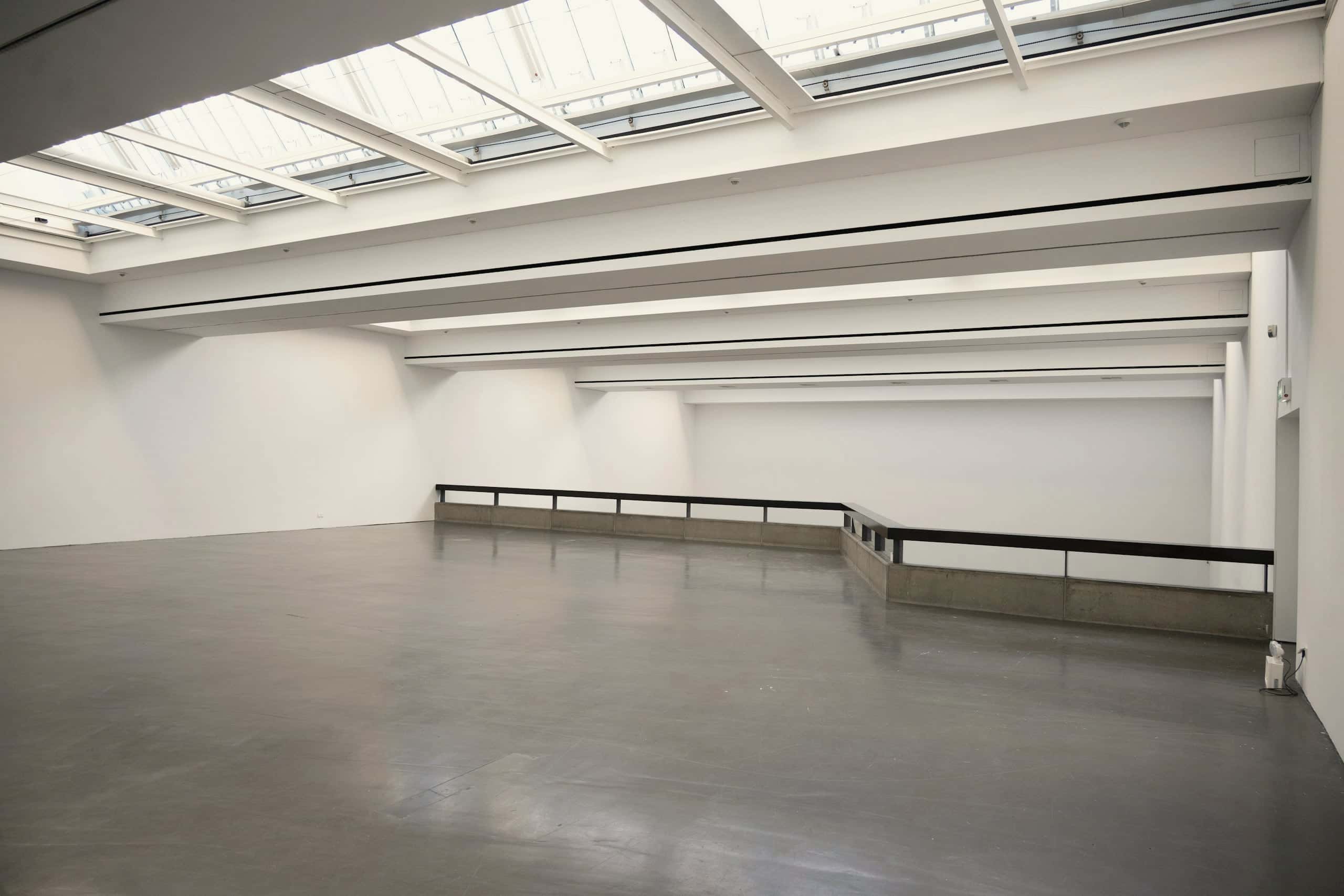 A long white gallery under a continuous run of rooflights, empty but for a handrail.