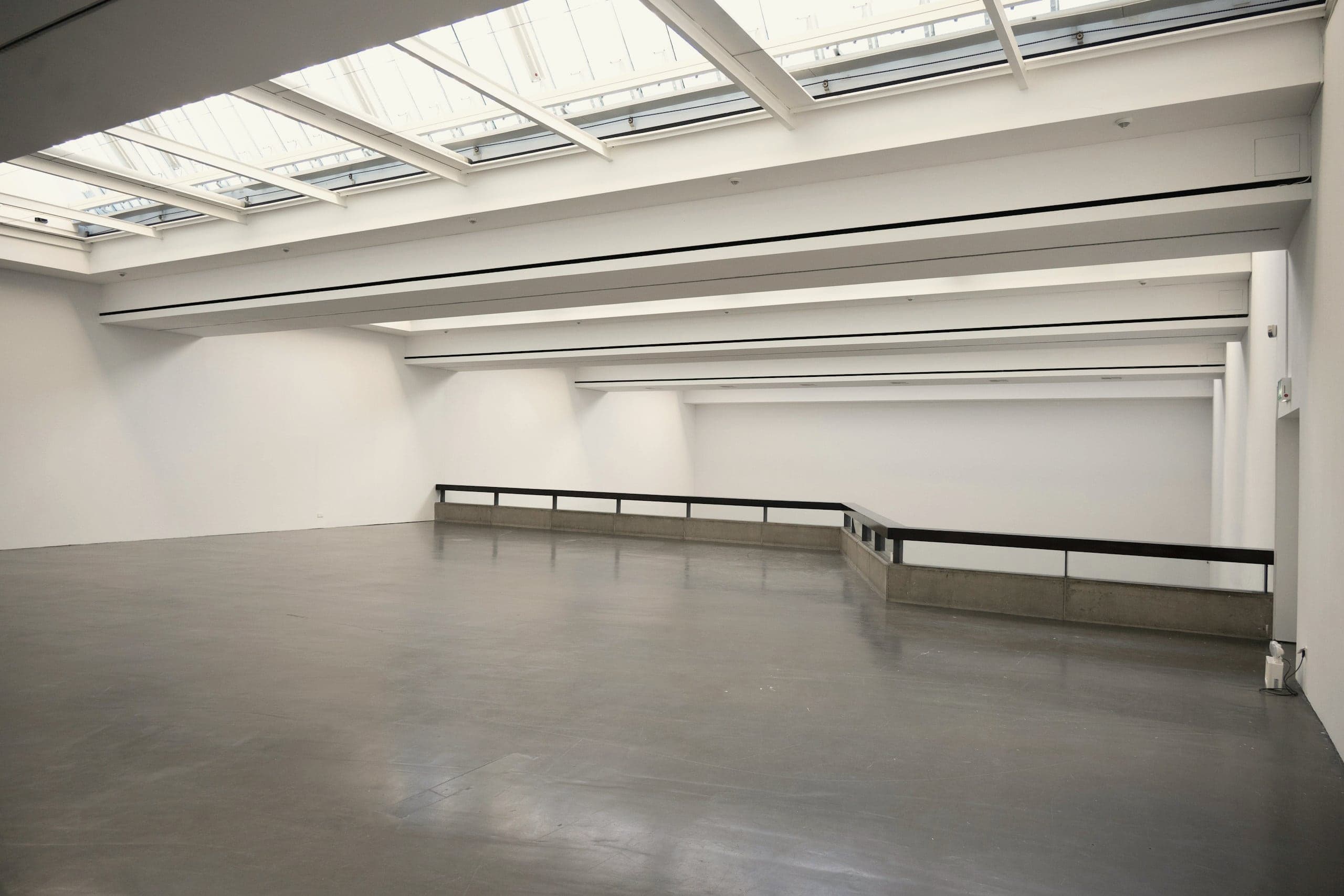 A long white gallery under a continuous run of rooflights, empty but for a handrail.