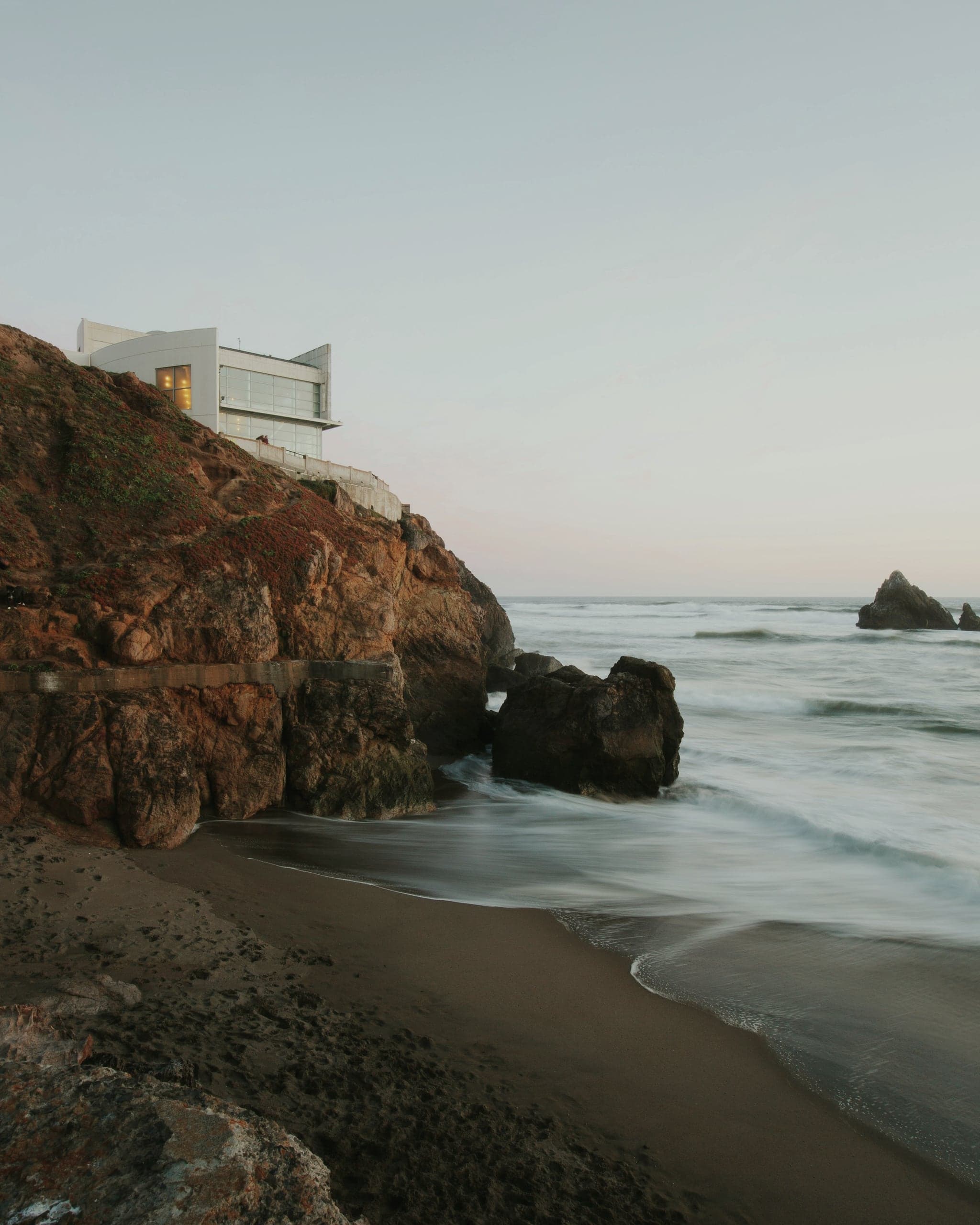 A house perched on a red rock cliff above a beach, the waves softened by long exposure.