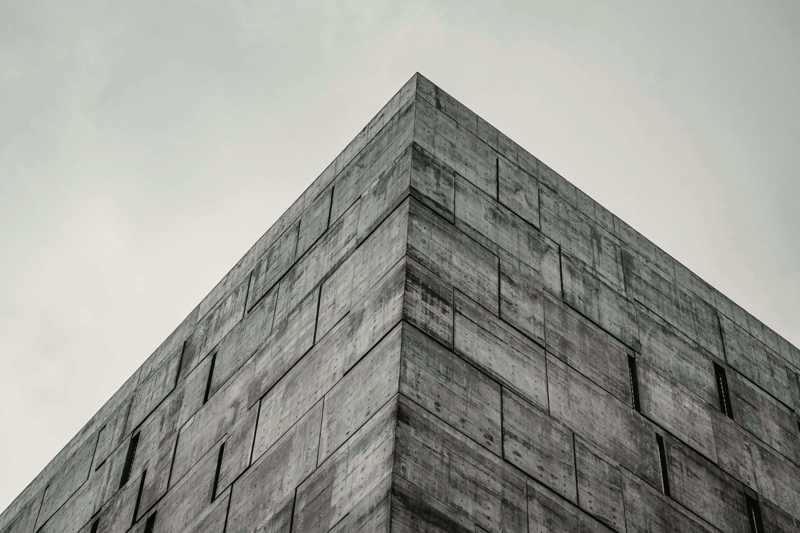 Corner of a board-marked concrete volume rising against an overcast sky, narrow window slots set flush in the wall.