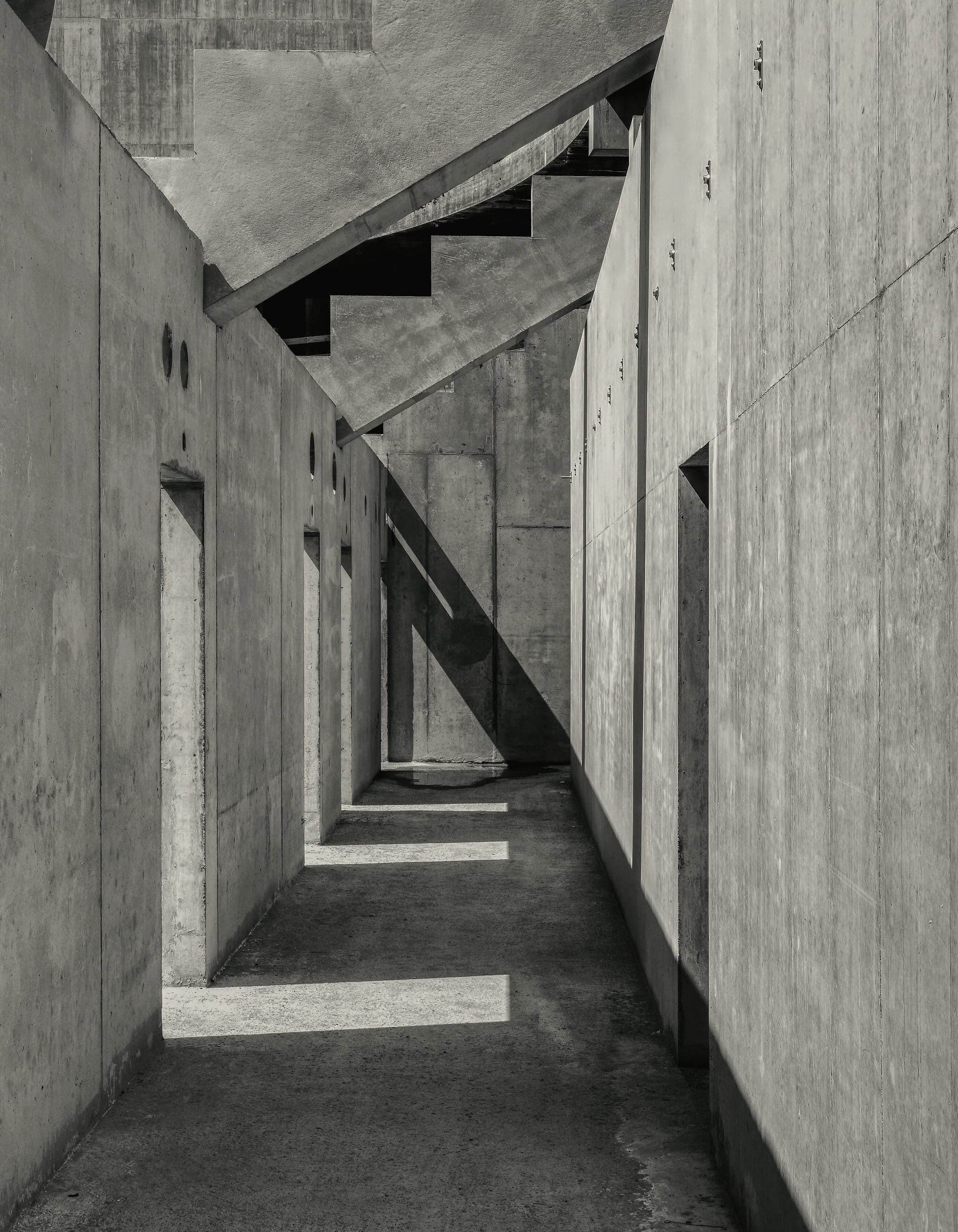 A narrow concrete passage with stairs overhead, lit in bands from one side.