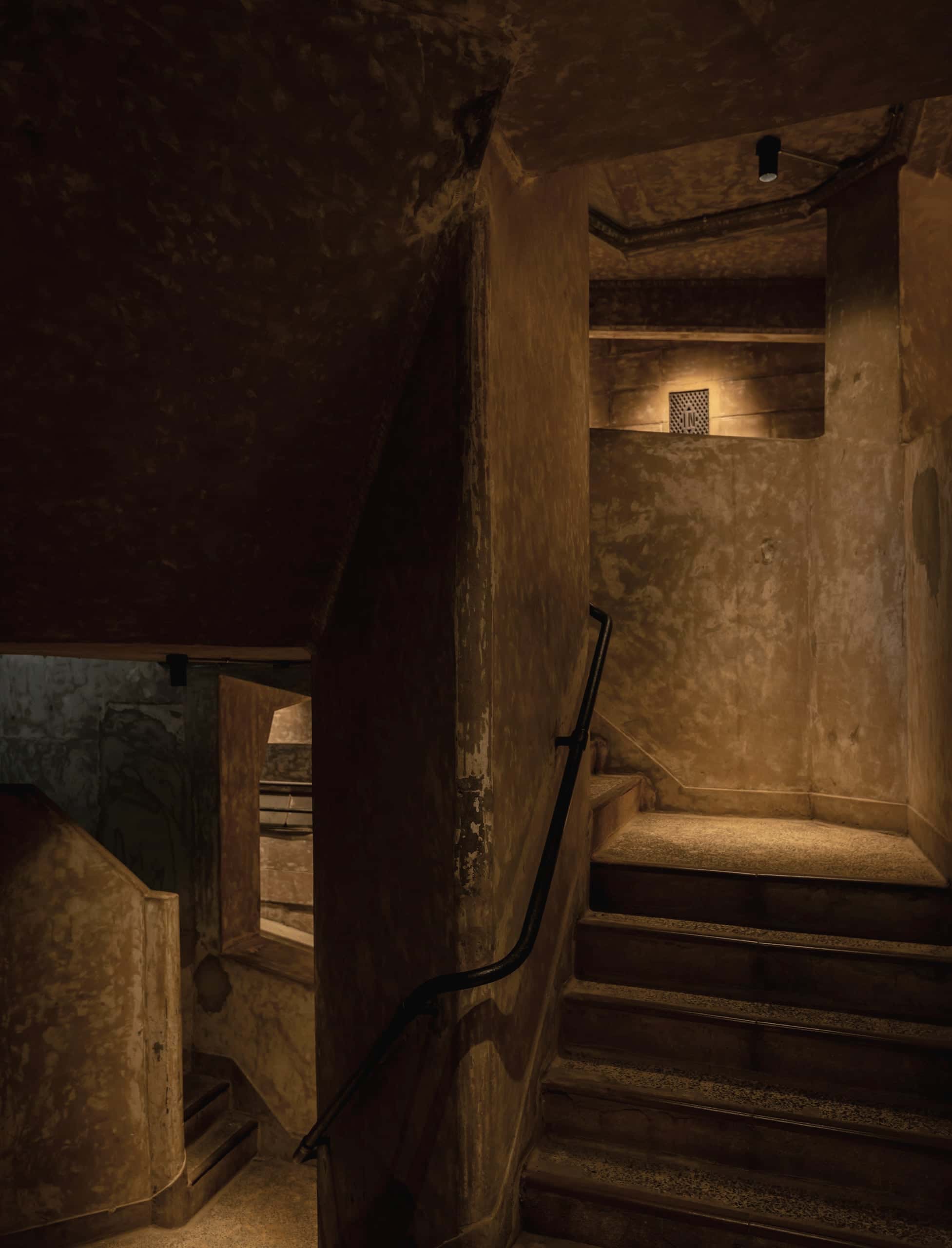 A narrow stone stair lit warm at the top of a dark stairwell.